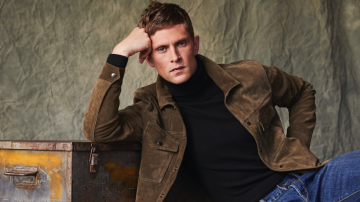 This image shows a male model posing with his head leaned against his hand in a brown jacket, black turtleneck and blue jeans.